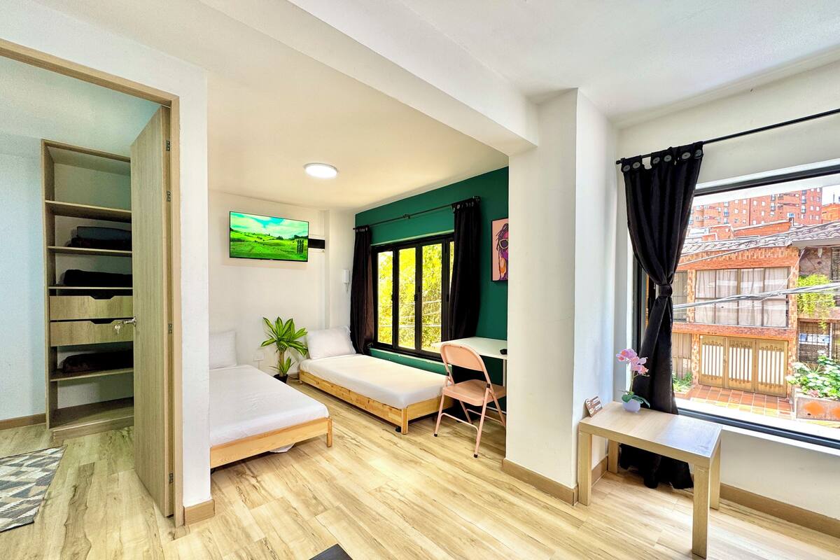 Quarto com cama, escrivaninha, cadeira, televisão e janela com vista para edifícios.