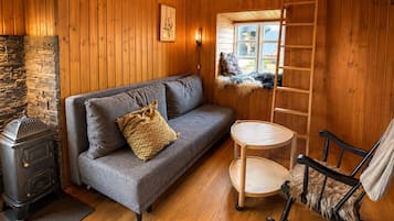 A cozy cabin interior with a stone fireplace, a gray sofa with a patterned pillow, a wooden chair, and a ladder to a loft.