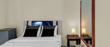 Habitación de hotel con cama, mesita de noche con lámpara, una pequeña mesa y un espejo grande.