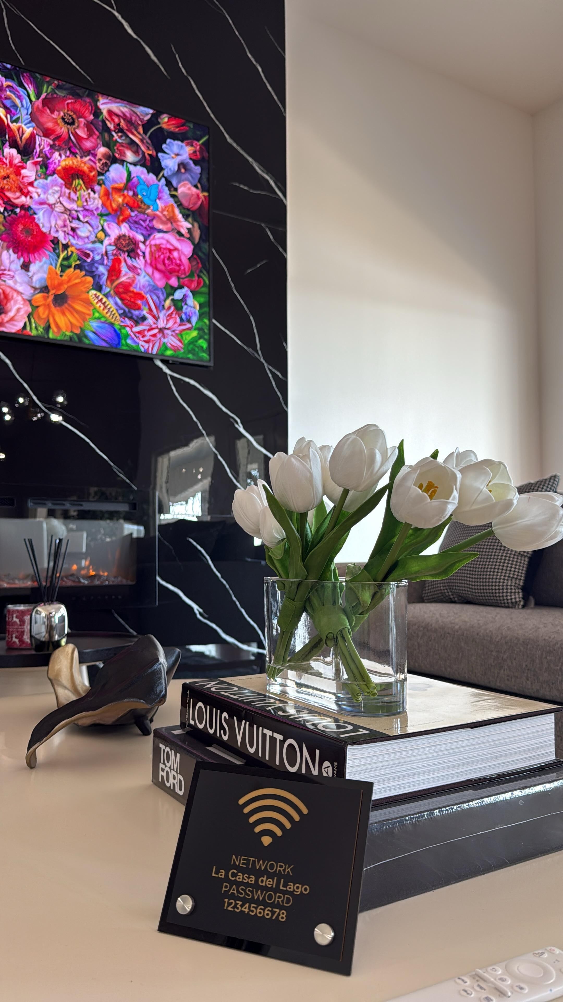 Ein modernes Wohnzimmer mit einem Flachbildfernseher, einer Vase mit weißen Tulpen, einem Buch mit dem Titel 'LOUIS VUITTON' und einem Schild mit einem WLAN-Passwort.