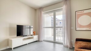 Ein modernes Wohnzimmer mit einem Flachbildfernseher auf einer weißen Konsole, einem braunen Sofa und einer großen Glasschiebetür, die zu einem Außenbalkon führt.