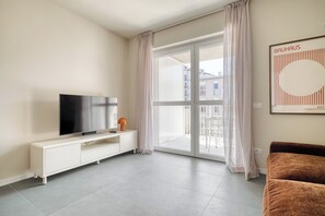 A modern living room with a flat-screen TV on a white console, a brown sofa, and a large sliding glass door leading to an outdoor balcony. - New apt terrace, parking, garden, metro in 2 min (Milan)