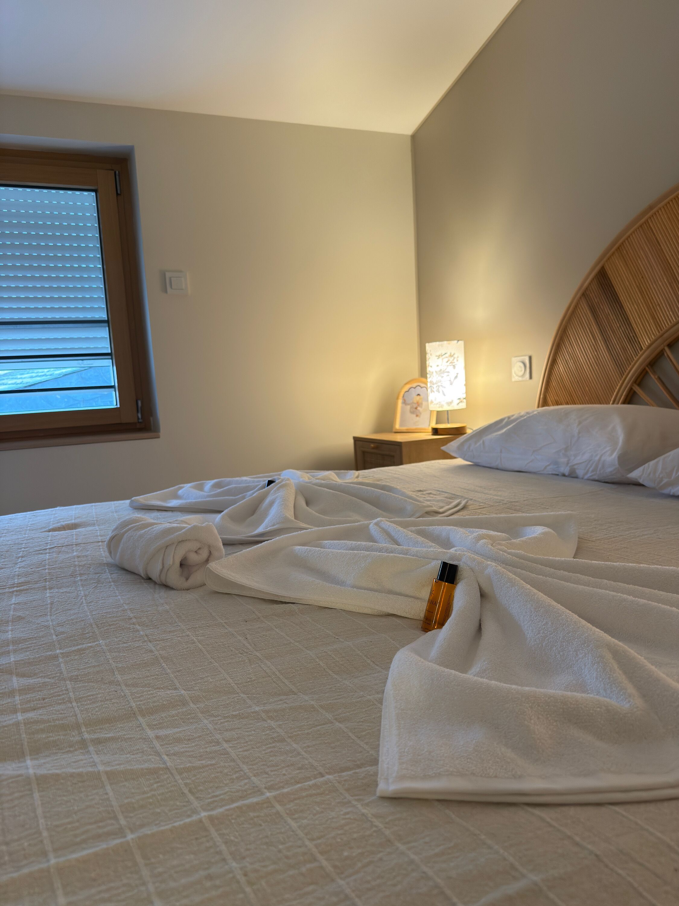 Un lit bien fait, avec une couette blanche, une lampe de chevet et une fenêtre avec des stores.
