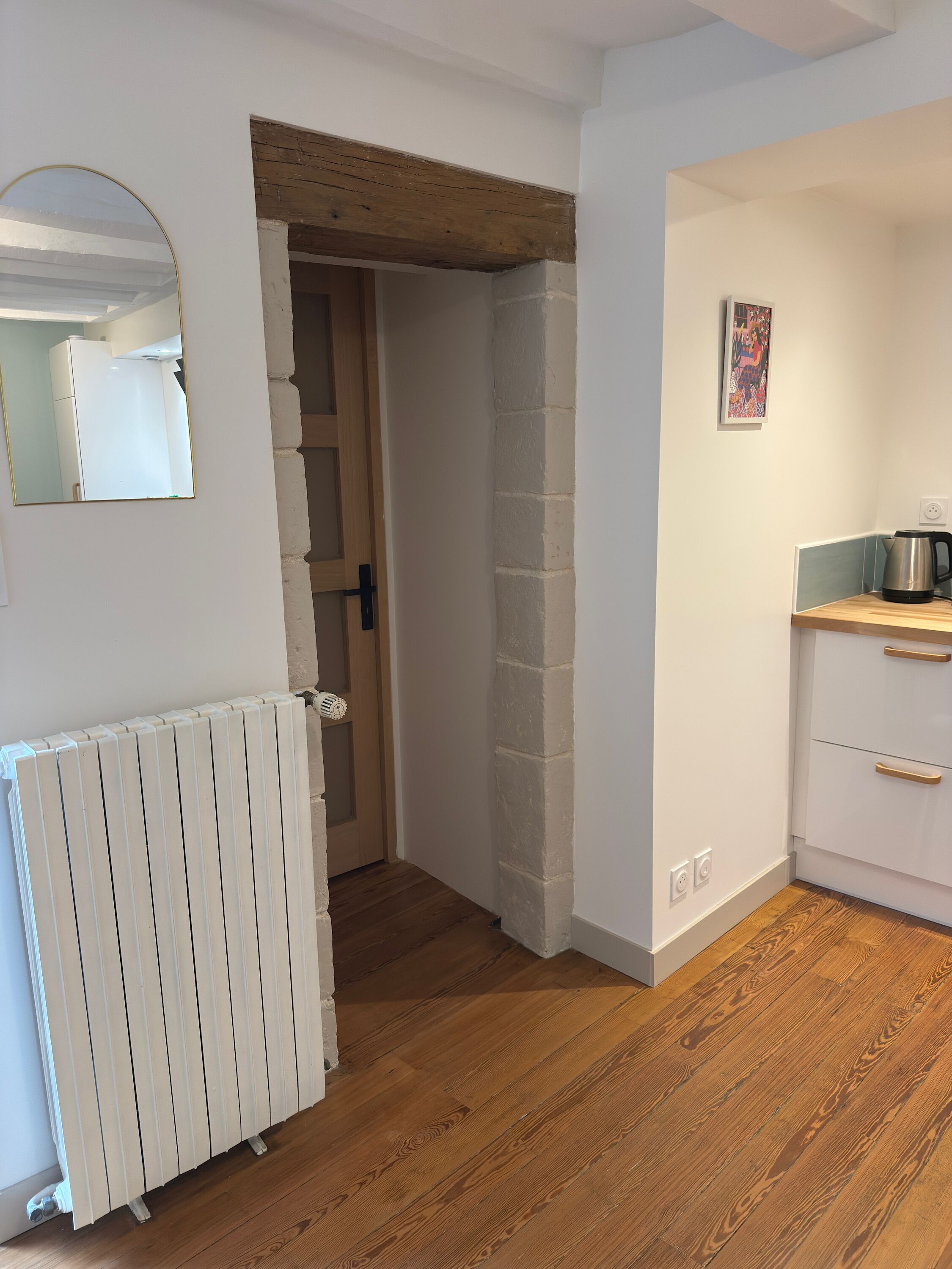 A room with a radiator, a wooden floor, a kitchen area with a kettle, and a mirror on the wall.