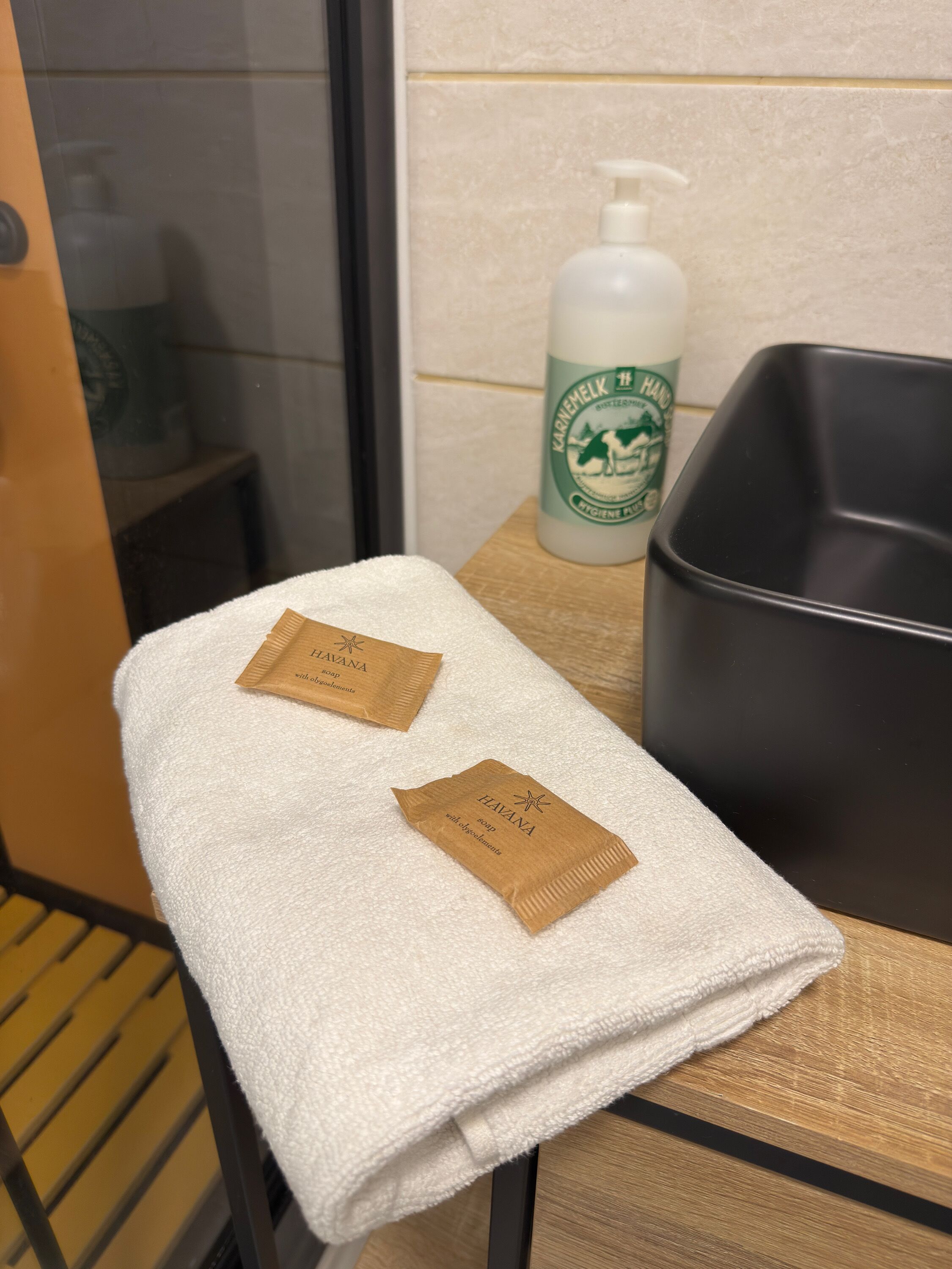 Een badkamer met een witte handdoek, twee zeepzakjes, een handzeepfles en een zwart wastafelbakje.