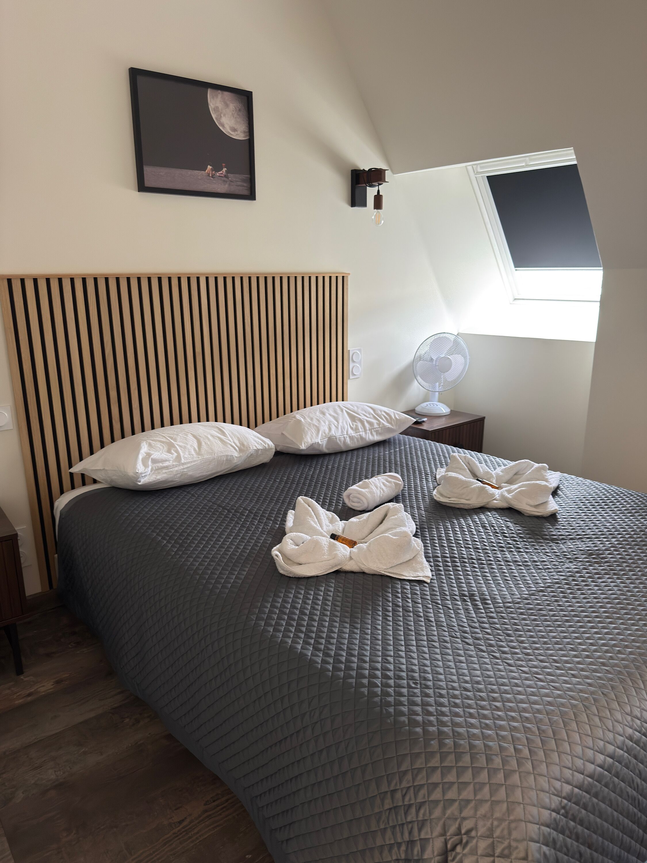A bedroom with a sloped ceiling, a bed with a quilted grey bedspread, white pillows, and a towel arrangement.
