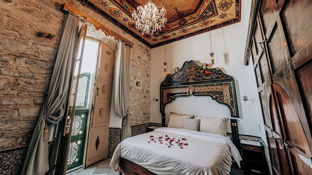 Ein Zimmer mit einer Steinwand, einem hölzernen Bett mit einem dekorativen Kopfteil, einer Chandelierleuchtre und einer grünen Tür.