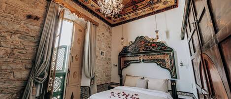Una habitación con pared de piedra, cama de madera con cabecera decorativa, candelabro y puerta verde.