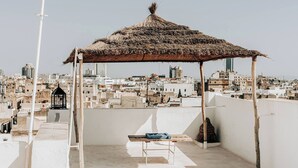 Eine Dachterrasse mit einer strohgedeckten Struktur, einer Bank und Blick auf eine Stadtlandschaft.