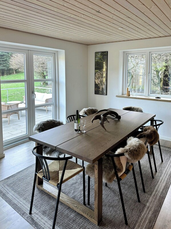 Un coin repas avec une table en bois, des chaises et une vue sur un jardin grâce à de grandes fenêtres.