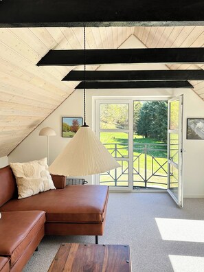 Een kamer op zolder met een bruine leren bank, een houten bijzettafeltje, een hanglamp en uitzicht op een groen landschap door een glazen deur.
