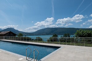 Ein Schwimmbecken mit Metallgeländern, freie Sicht auf einen See und Berge, sowie ein Gebäude mit Satteldach.