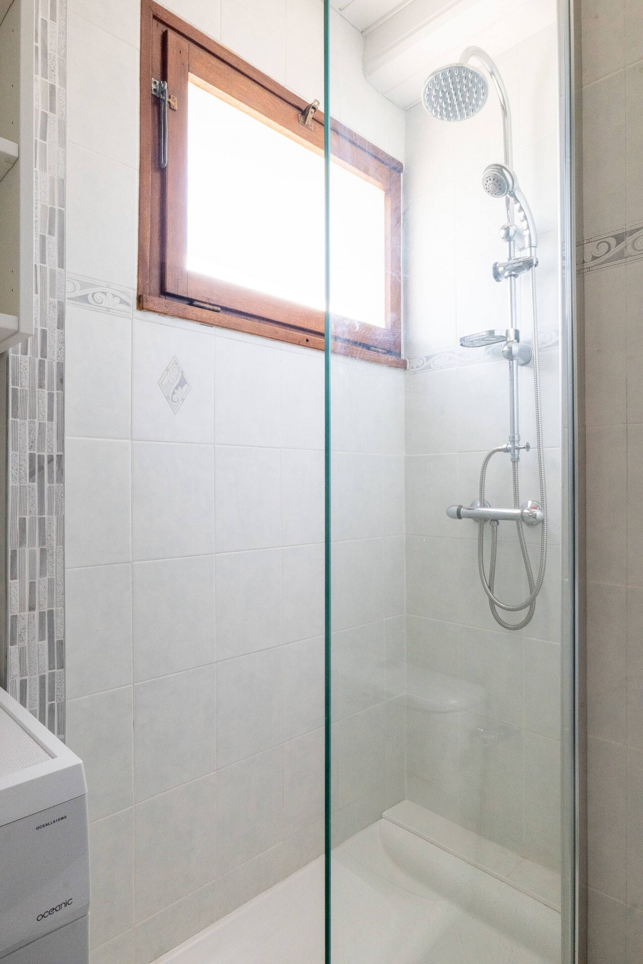 Ducha con paredes de vidrio, un cabezal de ducha de mano y una ventana en lo alto, además de un revestimiento de azulejos.