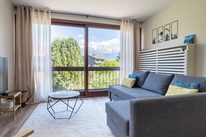 Ein modernes Wohnzimmer mit einer Couch, einem Glastisch und Blick auf Berge durch große Fenster.