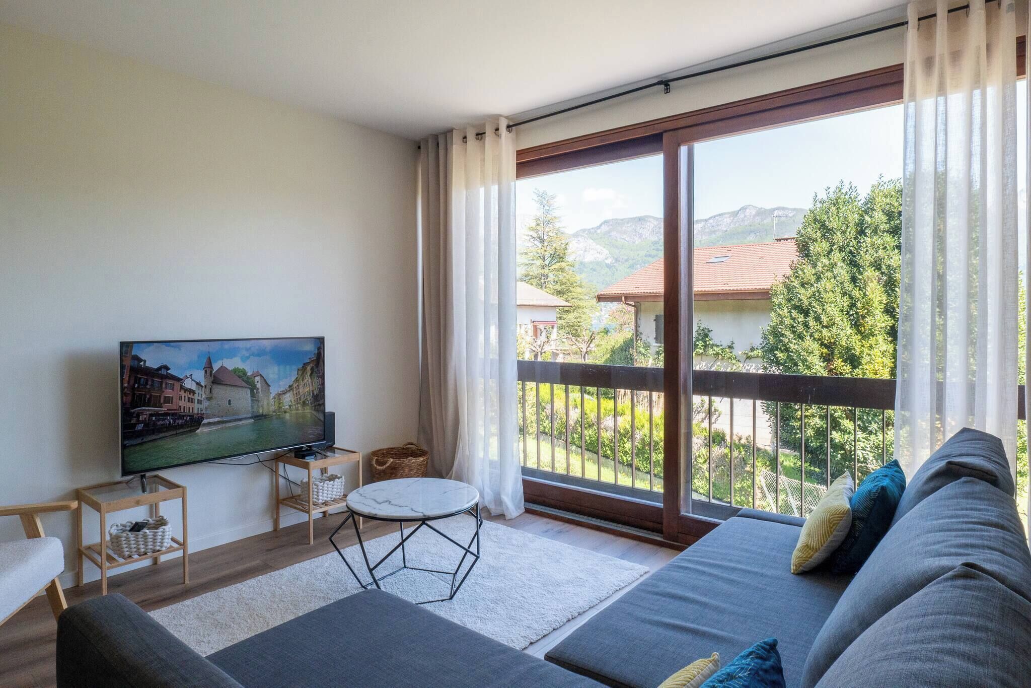 A modern living room with a large flat-screen TV, a sofa, a small round table, and a balcony with a view of a mountain and buildings.