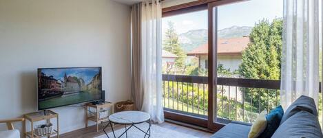 A modern living room with a large flat-screen TV, a sofa, a small round table, and a balcony with a view of a mountain and buildings.