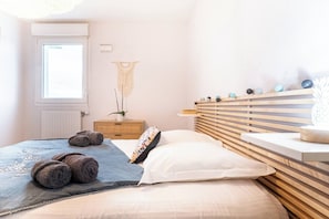 Ein Schlafzimmer mit Bett, Kissen, einem Nachttisch, einem an der Wand montierten Regal mit Dekoration und einem Wandbehang aus Makramee sowie einem Fenster.