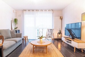Ein modernes Wohnzimmer mit einer Couch, einem runden Couchtisch, einem Fernseher und einem großen Fenster mit durchsichtigen Vorhängen.