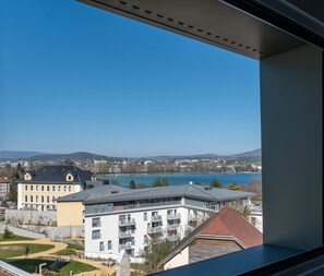 Blick aus einem Fenster auf eine Stadtlandschaft mit Gebäuden, einem See und fernen Hügeln.