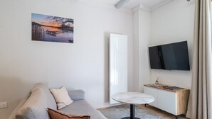 Ein modernes Wohnzimmer mit einer Couch, einem kleinen runden Tisch, einem Flachbildfernseher und einem Gemälde, das einen Sonnenuntergang zeigt.