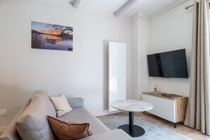 Ein modernes Wohnzimmer mit einer Couch, einem kleinen runden Tisch, einem Flachbildfernseher und einem Gemälde, das einen Sonnenuntergang zeigt.
