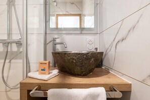 A bathroom with a marble wall, a wooden vanity, a large stone sink, a towel, and a welcome box. - - (Annecy)