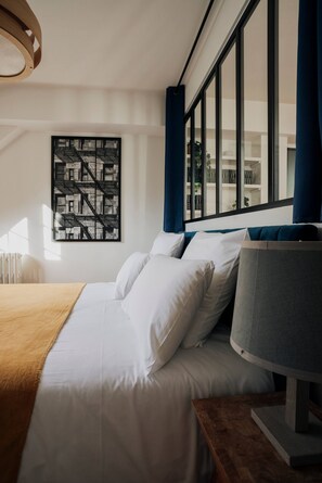 Ein modernes Hotelzimmer mit einem großen Bett, einem Beistelltisch, einer Wandlampe, einem Stadtbild als Kunstwerk im Bilderrahmen und Blick ins Freie durch große Fenster.