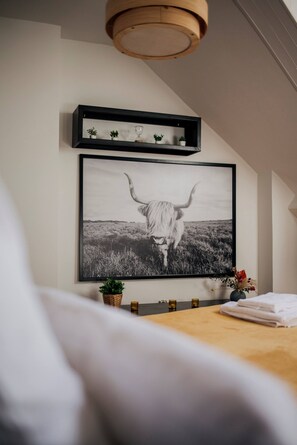 Ein gerahmtes Schwarz-Weiß-Foto einer Kuh auf einer Wiese, ein Regal mit kleinen Pflanzen und eine Vase mit Blumen auf einem Nachttisch.