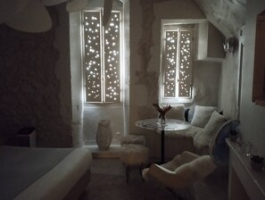 Ein Zimmer mit einem Bett, einem runden Tisch mit einer Vase, zwei Fenstern mit Lichtmustern, einem Sessel und einem Ventilator.