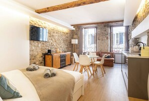 Ein gemütliches Zimmer mit Bett, Essbereich, Küche, Steinwänden und einem an der Wand befestigten Fernseher.