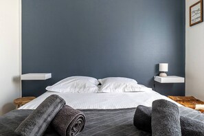 Un lit bien fait, avec du linge de lit blanc, une tête de lit bleu foncé et une petite table de chevet surmontée d’une lampe.