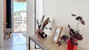 En gang med en glasdør, der fører ud til en balkon, en lille skammel, et indrammet billede og en rød vase med blade.