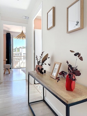 Ein Flur mit einer Glastür, die auf einen Balkon führt, einem kleinen Hocker, einem gerahmten Bild und einer roten Vase mit Blättern.