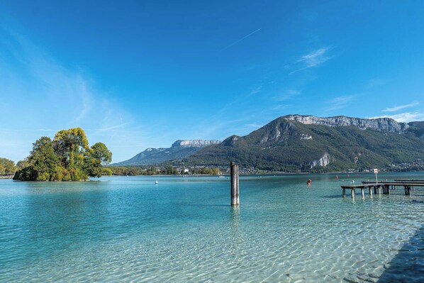 Un lac aux eaux limpides, avec une jetée en bois, une petite île parsemée d’arbres et des montagnes en arrière-plan.