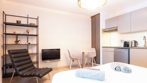 Una habitación de hotel moderna con cama, escritorio, silla, televisor, estantería y kitchenette.