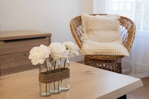Ein Holztisch mit drei Glasvasen, gefüllt mit weißen Blumen, ein Korbstuhl mit Kissen und ein Nachttisch im Hintergrund.