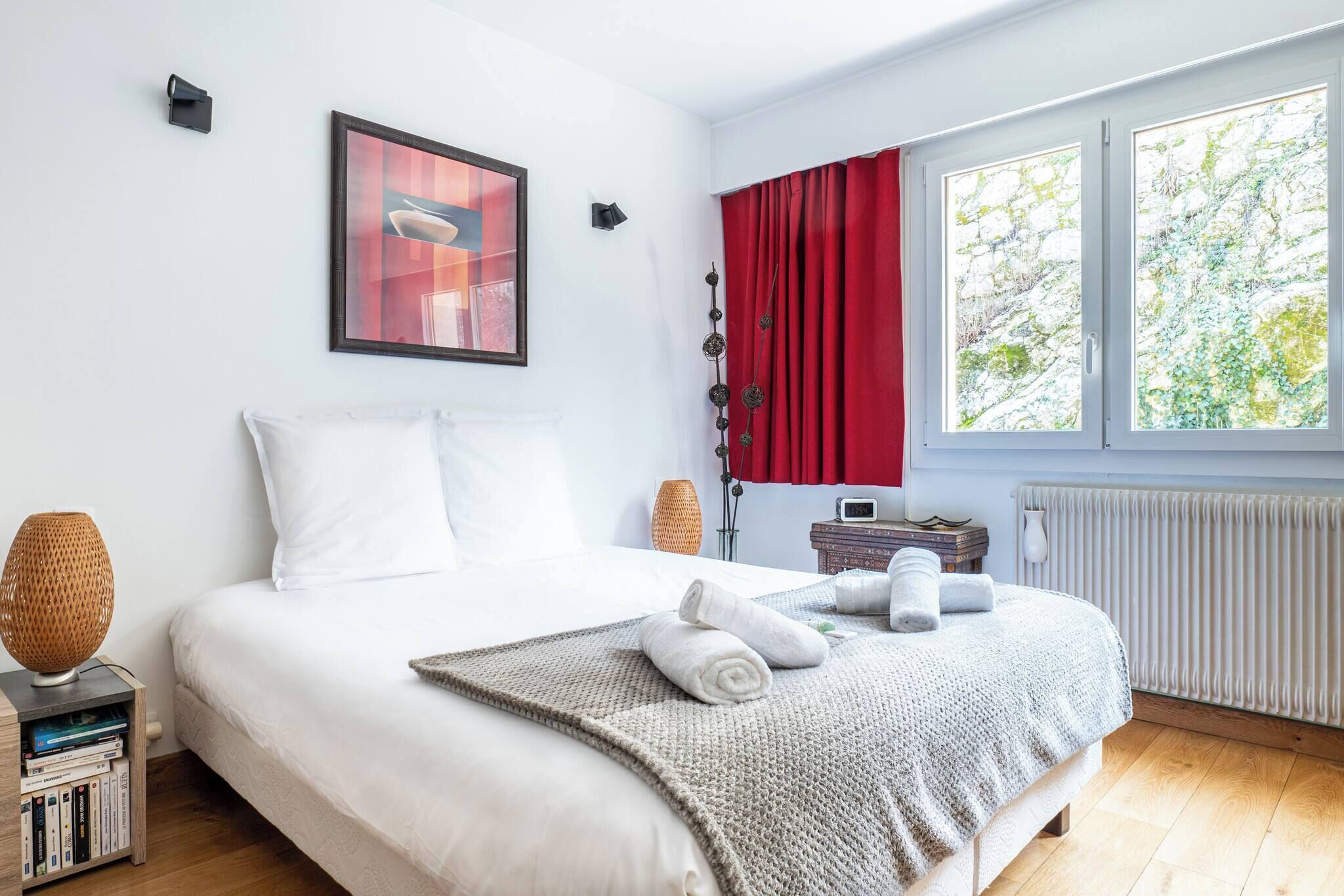 Ein ordentlich bezogenes Bett mit weißen Laken, eine Nachttischlampe und ein kleiner Tisch mit Büchern und einer Vase.
