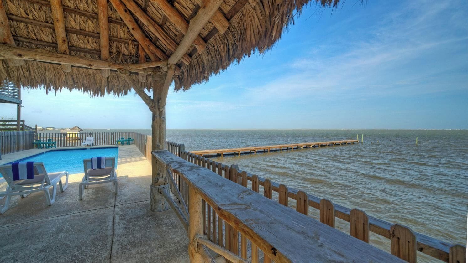 Un área de piscina con sillones reclinables, una terraza de madera y vista al agua.