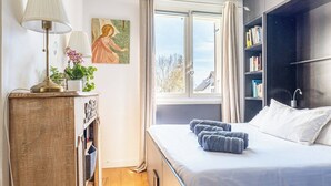 Ett sovrum med en säng, ett sängbord med en lampa, ett fönster med gardiner, en bokhylla och en tavla på väggen.