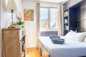 Ein Schlafzimmer mit Bett, Nachttisch mit Lampe, Fenster mit Vorhängen, Bücherregal und einem Bild an der Wand.