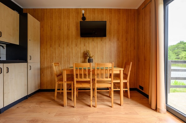 Una habitación con paneles de madera, una mesa de comedor y sillas, un televisor de pantalla plana montado en la pared y una ventana con vistas a la vegetación.