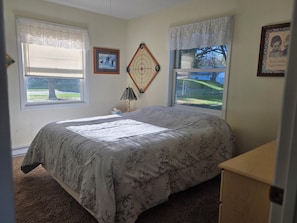 Une chambre à coucher avec un lit, deux fenêtres, une horloge, une lampe et un tableau encadré au mur.