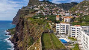 A coastal town with terraced houses, a modern building with a pool, and a steep cliff overlooking the sea. - Casa do Xano II by LovelyStay (Câmara De Lobos)
