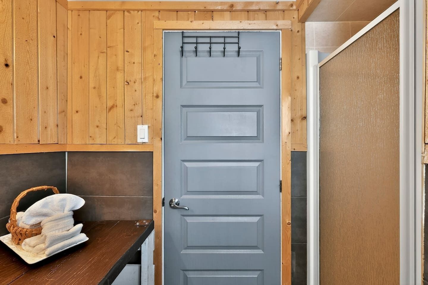Une pièce aux murs lambrissés, avec une porte grise, un mur recouvert de carreaux et un panier rempli de serviettes posé sur une table en bois.