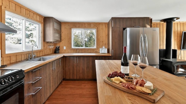 Une cuisine moderne avec des meubles en bois, un four encastré, une grande fenêtre et un plan de travail en bois agrémenté d’une planche à fromage.
