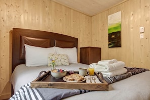 Quarto de hotel com cabeceira de madeira, cama com roupa de cama branca, bandeja com itens de café da manhã e um quadro na parede.