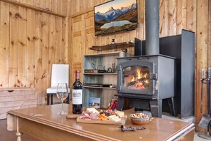 Interior de madeira com lareira, uma garrafa de vinho, queijo e um livro sobre uma mesa.