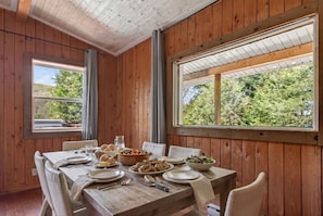 Uma área de refeições com mesa de madeira posta para quatro pessoas, rodeada por paredes de madeira e grandes janelas.