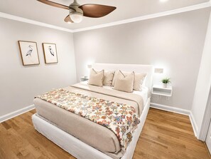 Une chambre à coucher avec un lit, un ventilateur de plafond, deux tableaux accrochés au mur, des tables de chevet avec des lampes et un plancher en bois.