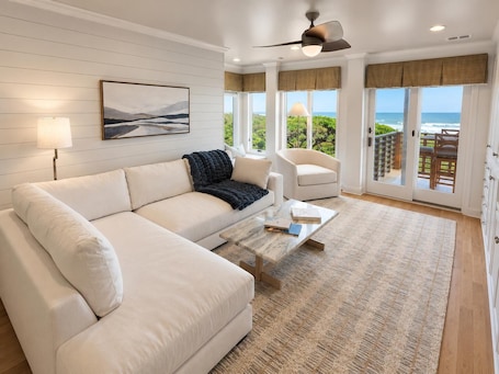 A modern living room with a white sofa, a coffee table, a ceiling fan, and a view of the ocean through large windows.. Ocean View Escape | Dual King Suites | East Beach